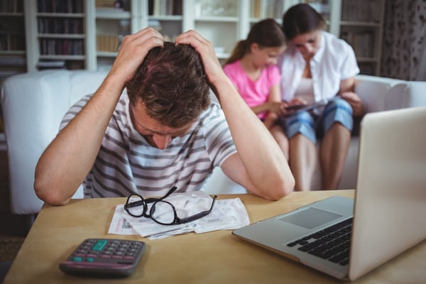 Worried man sitting at table with bills and laptop while his wife and daughter sitting on sofa-1